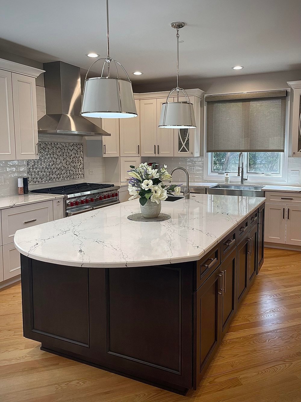 Spacious kitchen featuring white shaker cabinets and subway‑tile backsplash, a dark wood island topped with veined white quartz and a floral centerpiece, stainless‑steel range with mosaic tile accent, two pendant lights, a large window with a roller shade, and light hardwood floors.