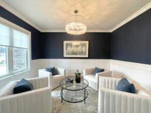 Services 5 Chic sitting area with four white upholstered armchairs arranged around a round glass‑top coffee table styled with a potted plant, set against navy blue walls above white wainscoting, under a crystal chandelier and abstract wall art.