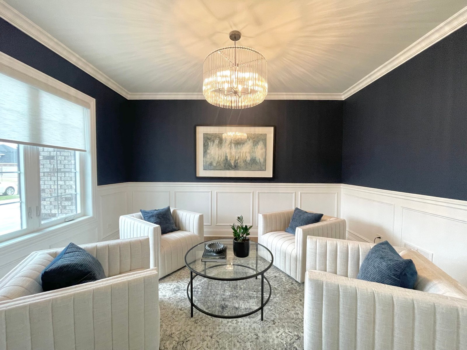 Chic sitting area with four white upholstered armchairs arranged around a round glass‑top coffee table styled with a potted plant, set against navy blue walls above white wainscoting, under a crystal chandelier and abstract wall art.