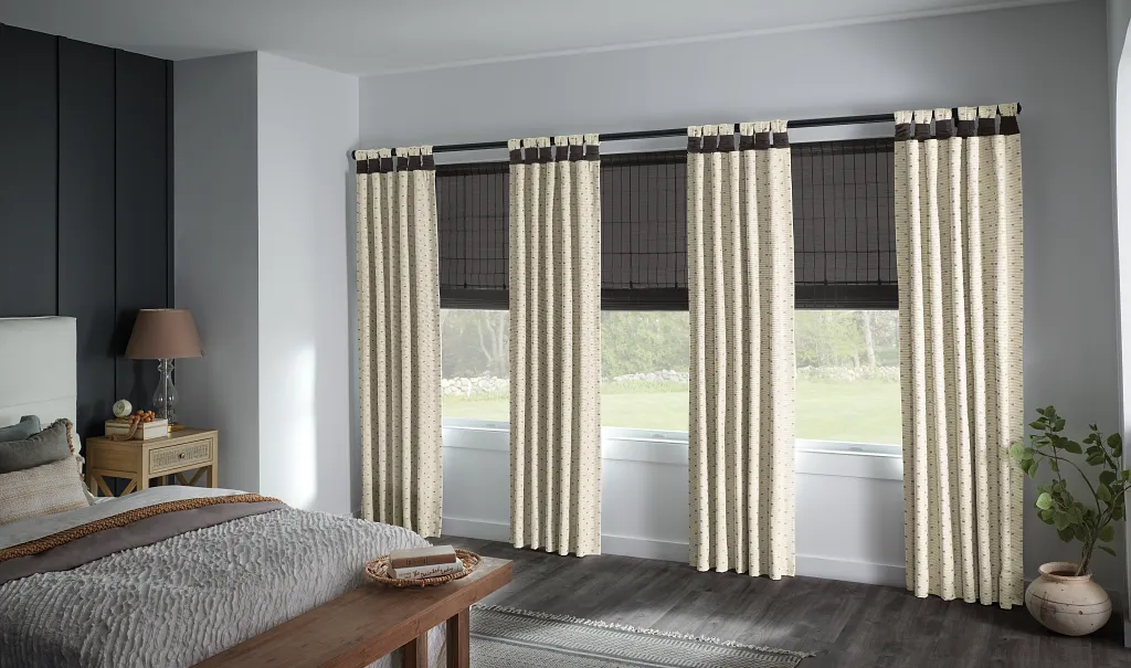 A soft, layered combination of Graber window treatments in a bedroom features blackout shades with tailored drapes. These custom window treatments offer style and light control while adding warmth and texture to the sleeping space.
