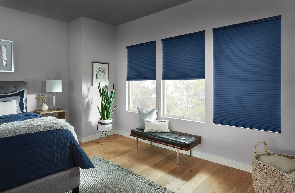 graber-custom-blinds-4 Modern bedroom featuring deep blue cellular shades on three large windows, paired with neutral gray walls, a bench, and minimalist decor.