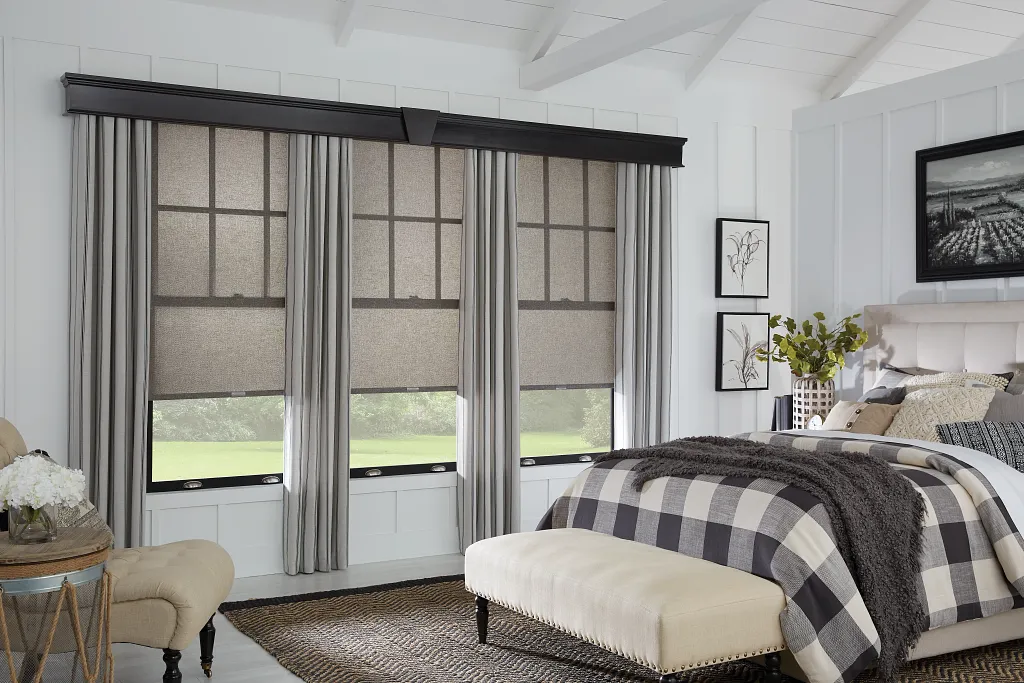 Elegant bedroom showcasing layered window treatments with light gray drapes and textured roller shades, framed by white walls and a vaulted ceiling.