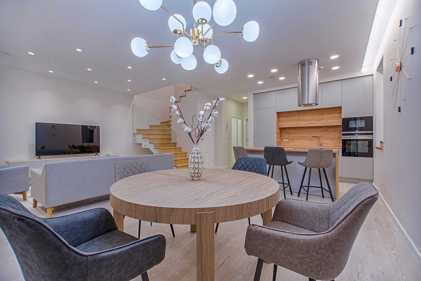 Modern open-concept living space featuring a round wood dining table with four upholstered chairs, a minimalist gray sofa, and a sleek kitchen with light gray cabinets and wood accents. A statement gold chandelier with globe lights hangs overhead, tying together this contemporary furniture style with clean interior decorating ideas and cohesive furniture styling.
