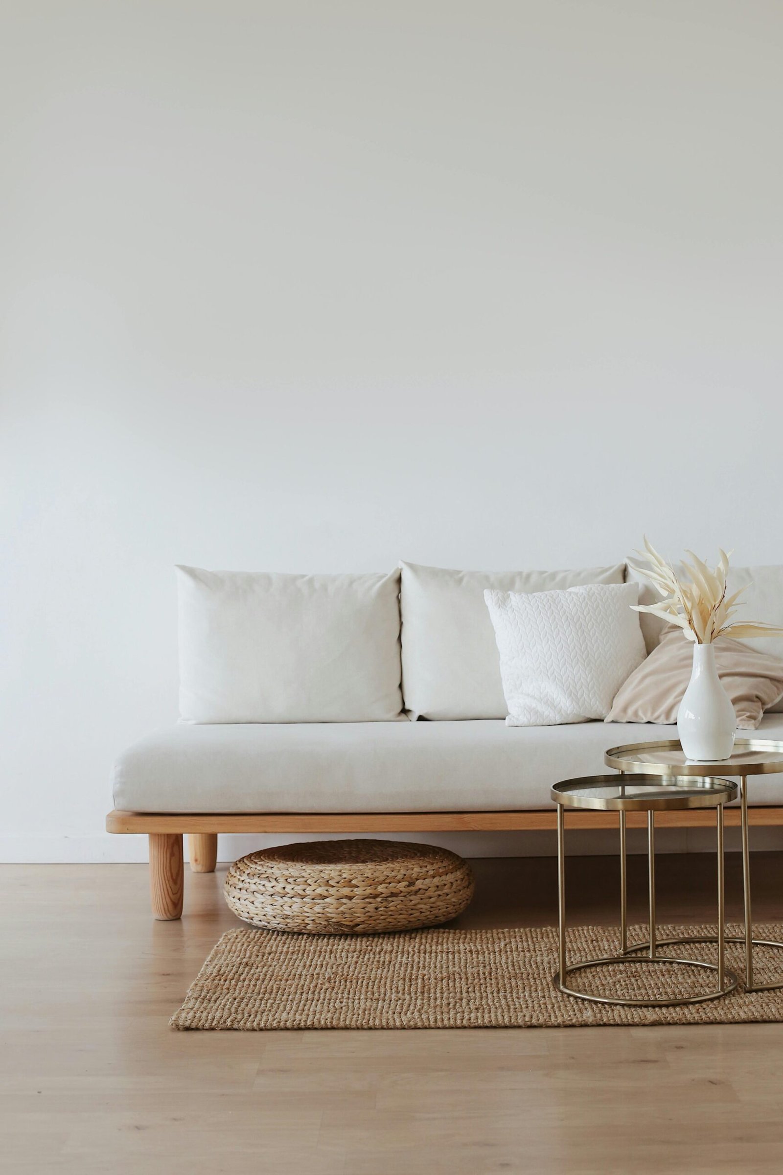 Minimalist living room featuring a light wood frame sofa with soft white cushions, a woven jute rug, and two modern brass side tables. A white ceramic vase with dried foliage adds a decorative touch, creating a warm and calming furniture styling aesthetic.