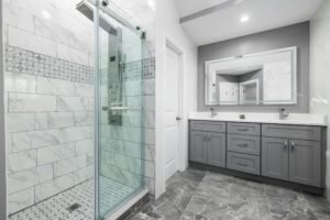 Furniture, Decor & Styling 4 Modern bathroom with a sleek walk-in glass shower featuring marble tiles and geometric accent bands. A large gray double vanity with chrome hardware and white countertops sits beneath a wide backlit mirror, combining contemporary design with functional elegance.