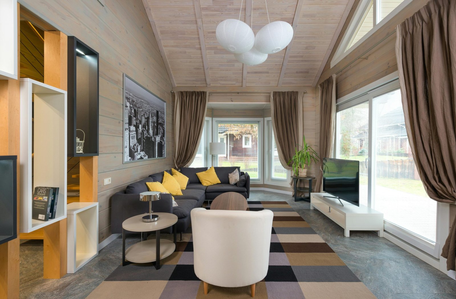 Cozy contemporary living room with vaulted wood-paneled ceiling, a large sectional sofa with yellow accent pillows, and full-length brown curtains. A modern white armchair and checkered area rug anchor the seating space, while a flat-screen TV sits on a low white stand beside expansive sliding glass doors. A black-and-white cityscape print and indoor plant complete the warm, welcoming design.