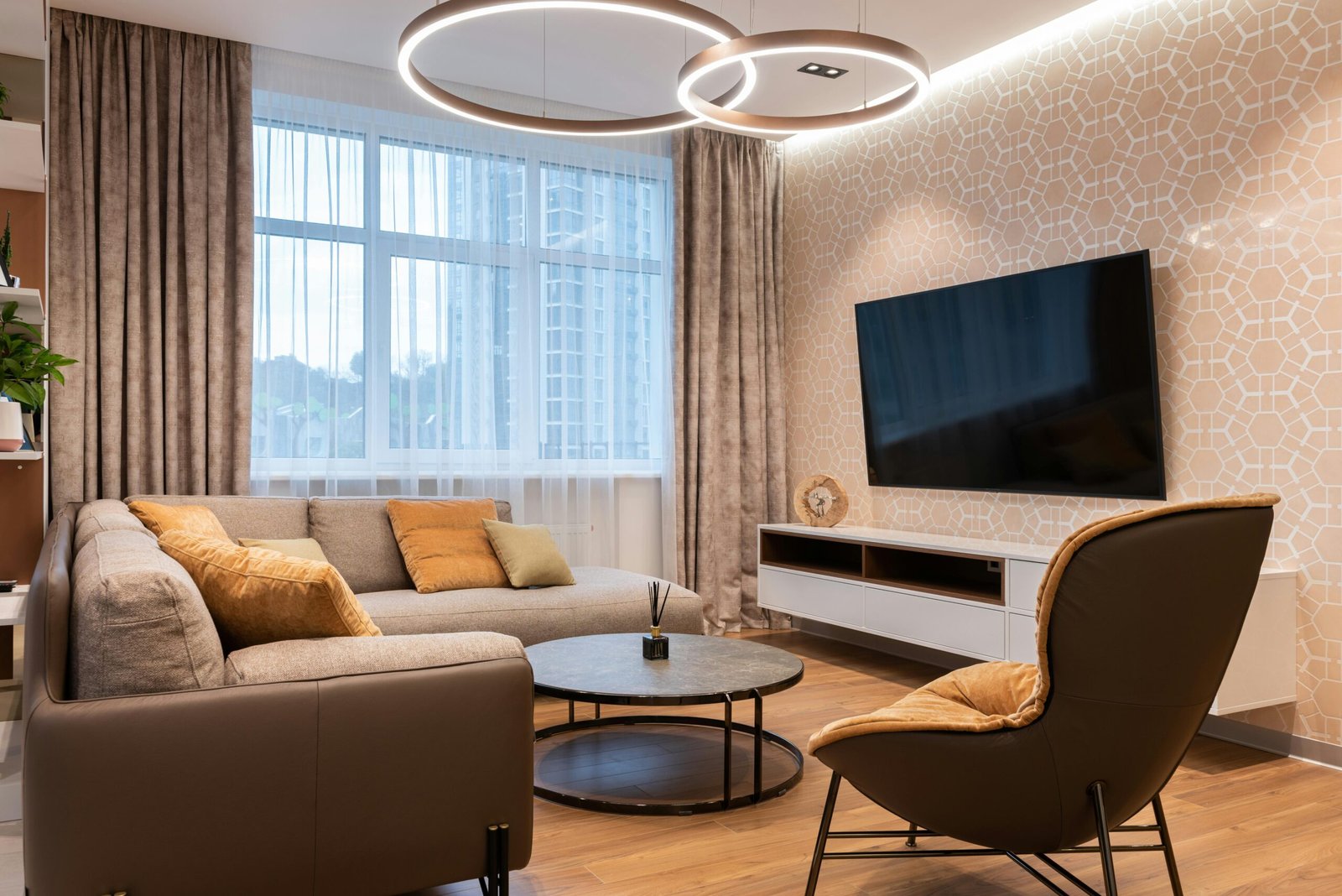 Cozy contemporary living room with vaulted wood-paneled ceiling, a large sectional sofa with yellow accent pillows, and full-length brown curtains. A modern white armchair and checkered area rug anchor the seating space, while a flat-screen TV sits on a low white stand beside expansive sliding glass doors. A black-and-white cityscape print and indoor plant complete the warm, welcoming design.