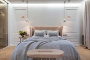 Furniture, Decor & Styling 7 Modern bedroom featuring a blush pink upholstered headboard and soft blue bedding with matching pillows. A pair of pendant lights—one silver and one rose gold - hang symmetrically above each side of the bed. A round bedside table holds a fresh floral arrangement, and a velvet bench sits at the foot of the bed. Decorative wall molding and soft, ambient lighting create a serene and sophisticated atmosphere.