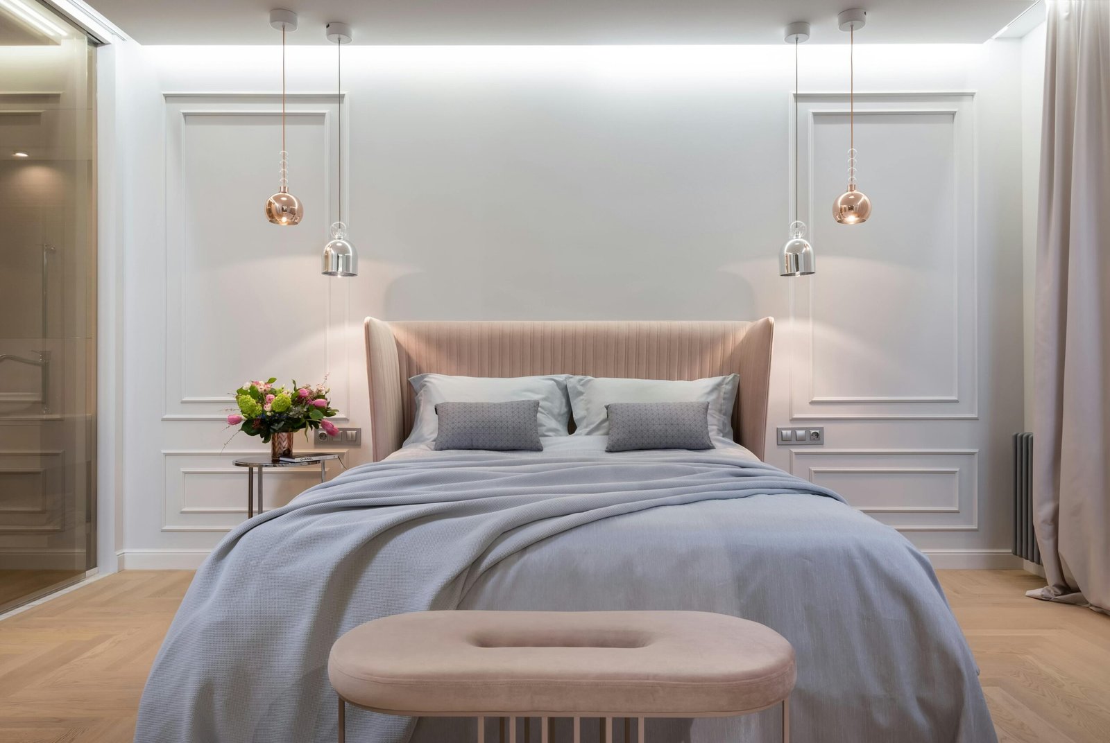 Modern bedroom featuring a blush pink upholstered headboard and soft blue bedding with matching pillows. A pair of pendant lights—one silver and one rose gold - hang symmetrically above each side of the bed. A round bedside table holds a fresh floral arrangement, and a velvet bench sits at the foot of the bed. Decorative wall molding and soft, ambient lighting create a serene and sophisticated atmosphere.