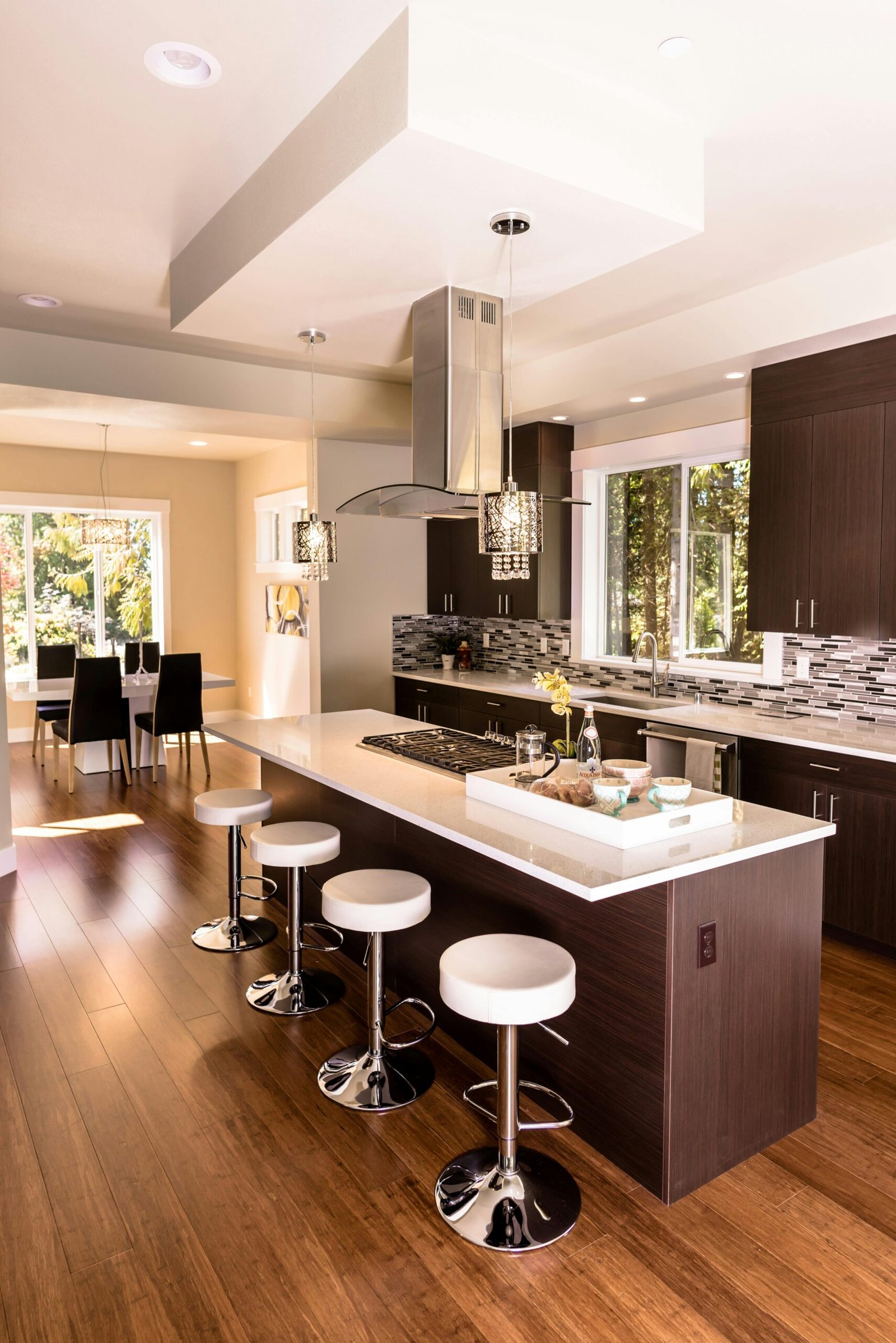 Contemporary kitchen with dark wood cabinetry, white quartz countertops, and a large island featuring a built-in gas cooktop and four white cushioned barstools. Pendant lights with crystal accents hang above the island, adding elegance. A mosaic tile backsplash complements the modern design, and natural light fills the space through large windows. In the background, a dining area with black chairs and a white table enhances the open-concept layout.