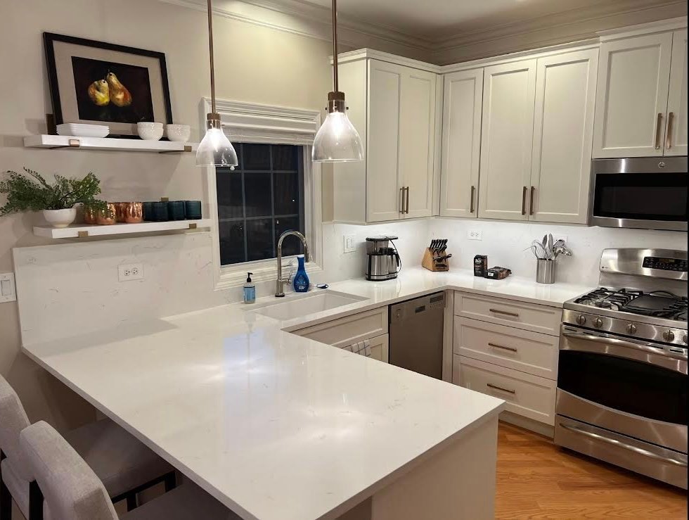 Kitchen with white shaker cabinets, marble‑effect quartz countertops, stainless‑steel range and microwave, glass pendant lights, floating shelves styled with decor, and light hardwood flooring.