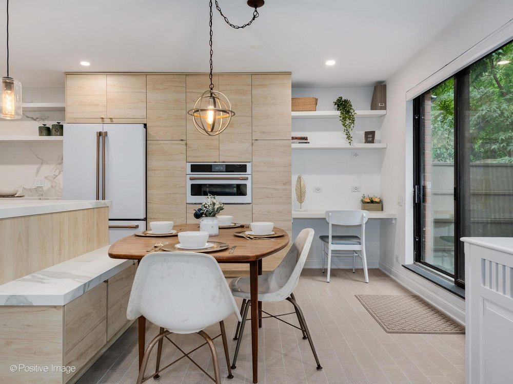 Bright open kitchen with light wood cabinets and white marble countertops, featuring a built‑in refrigerator and oven wall, a small round dining table set for four, a built‑in desk nook with shelving, and a sliding glass door overlooking greenery.
