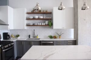 Kitchen Design 8 Modern kitchen with white upper and gray lower cabinets, marble‑veined quartz countertops, open wood shelving, subway tile backsplash, stainless‑steel range and sink, and chrome pendant lights.
