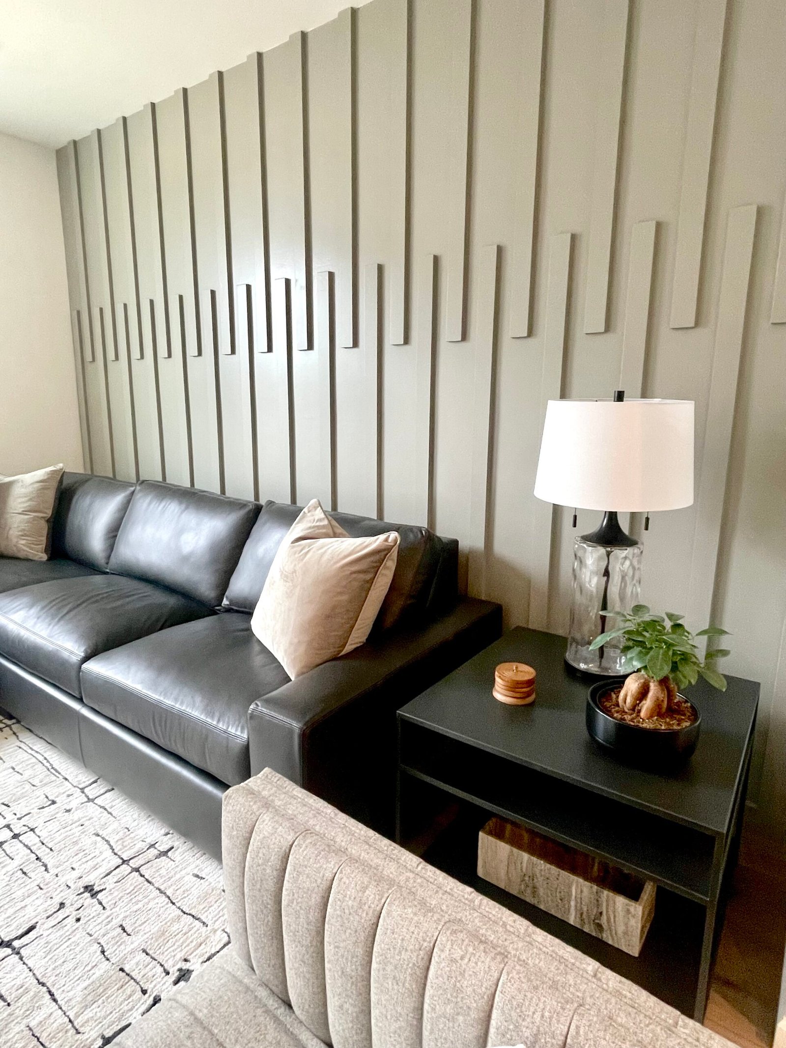 Black leather sofa with beige throw pillows against a grey vertical‑panel accent wall, beside a black side table topped with a glass‑based lamp and potted plant, with a portion of a light upholstered chair in the foreground.