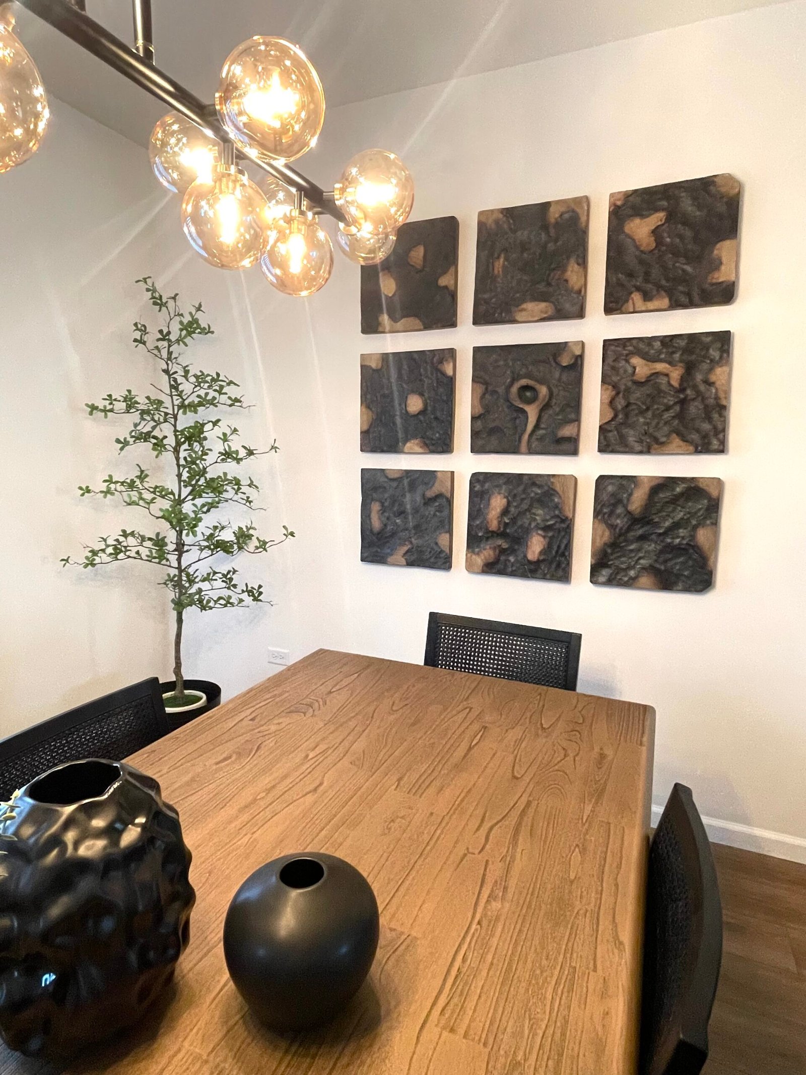 Contemporary dining area with a light wood table topped by two black vases, surrounded by black mesh chairs, illuminated by a multi‑bulb chandelier, accented by a tall potted plant and a 3×3 grid of textured wood wall art panels.