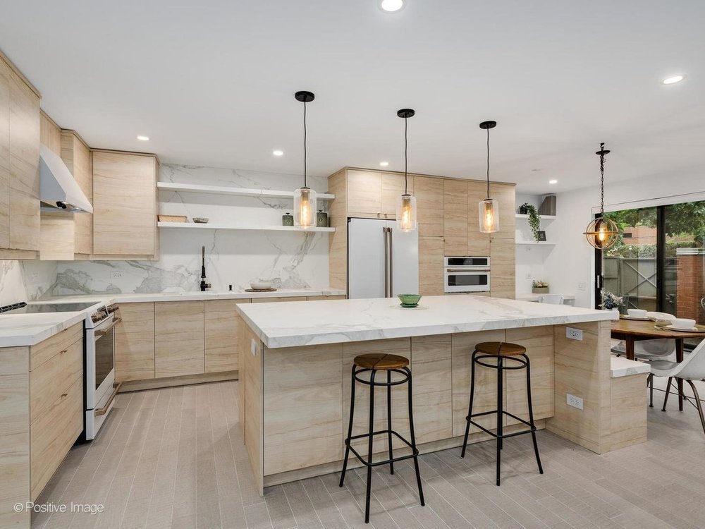 Open kitchen with light wood cabinetry and white marble countertops, featuring a large island with two stools, three glass pendant lights, built‑in stainless appliances, open shelves, and a sliding glass door to the backyard.