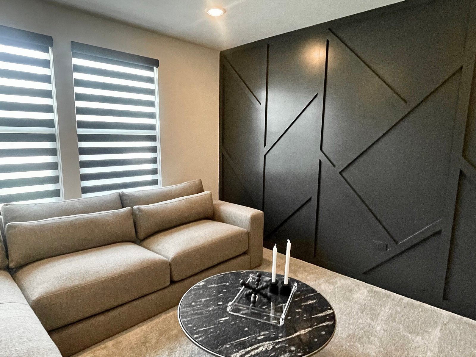 Light beige upholstered sectional sofa facing a matte black geometric accent wall with diagonal molding, zebra‑striped roller shades on twin windows, and a round black marble coffee table topped with candles on a light carpet.