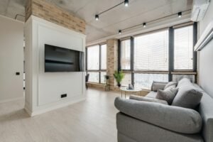 Window Treatments 8 Modern living room with floor-to-ceiling windows covered in light wood horizontal blinds, paired with a gray sectional sofa, exposed brick pillars, and a mounted flat-screen TV.