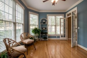 Window Treatments 9 Bright sunroom with white horizontal blinds on multiple windows, soft blue walls, natural wood floors, and two rattan accent chairs with beige cushions.