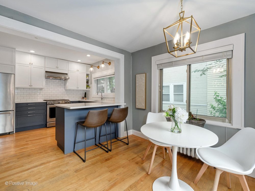 Open kitchen and breakfast nook featuring white upper cabinets, navy blue island with two brown bar stools, a round white tulip table with chairs, large window dressed in horizontal blinds, brass lantern pendant, and light hardwood floors.