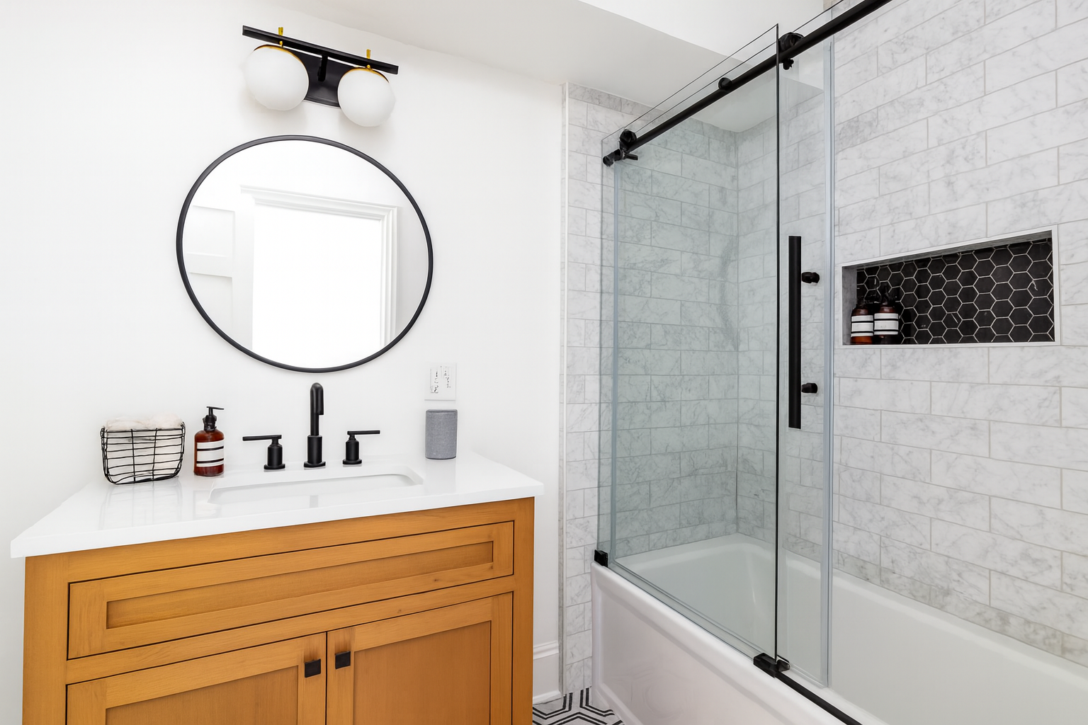 Modern bathroom with a warm wood vanity, black round mirror, and matte black fixtures. The space features a tub-shower combo with sliding glass doors, marble subway tile walls, and a recessed black hexagon tile niche holding toiletries.