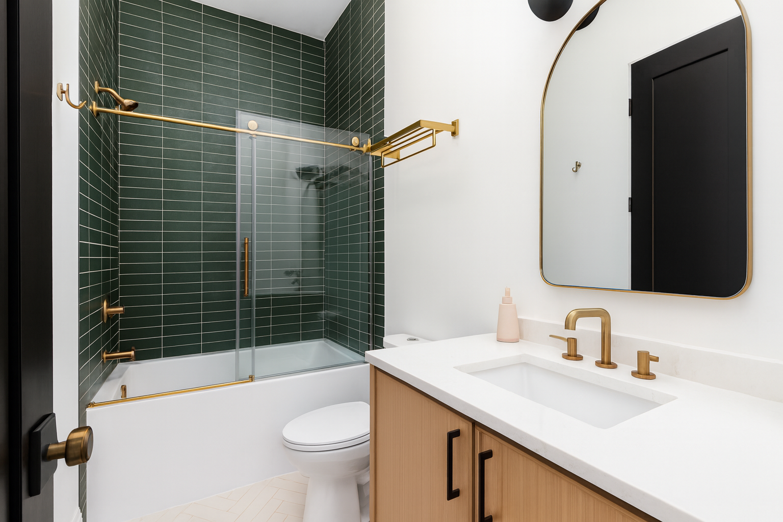 Contemporary bathroom with deep green subway tile shower walls accented by gold fixtures and hardware. A floating glass shower door complements the sleek wood vanity with black pulls, topped with a white countertop and gold faucet beneath an arched mirror.