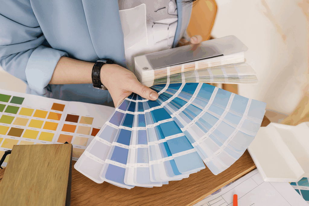 Closeup Of Architect Woman Choosing Samples Of Wall Paint Interior Designer Looking At Color Swatch For Creating Project House Renovation Architecture And Interior Design Concept Interior designer reviewing paint swatches, holding a fan deck of blue shades while other color samples and design materials spread across the desk. The scene highlights the process of choosing wall paint and creating a project concept.
