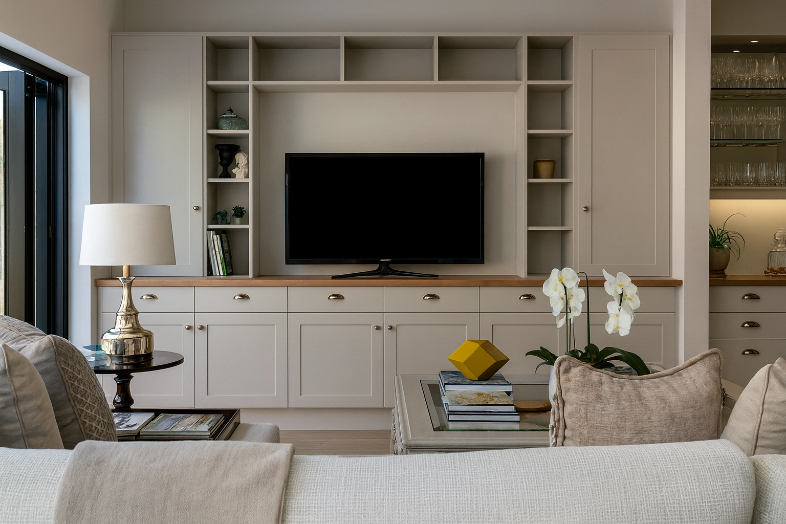 Modern living room with built-in cabinetry and shelving framing a flat-screen TV. Neutral tones, wood accents, and decorative objects like books, plants, and orchids create a balanced and inviting design.