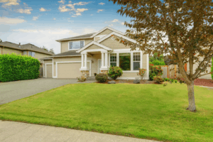 Interior & Exterior Paint Consultations 6 Two-story beige suburban home with white trim, stone accents, and a double garage. A neatly landscaped front yard with trees and shrubs complements the wide driveway and welcoming entryway.