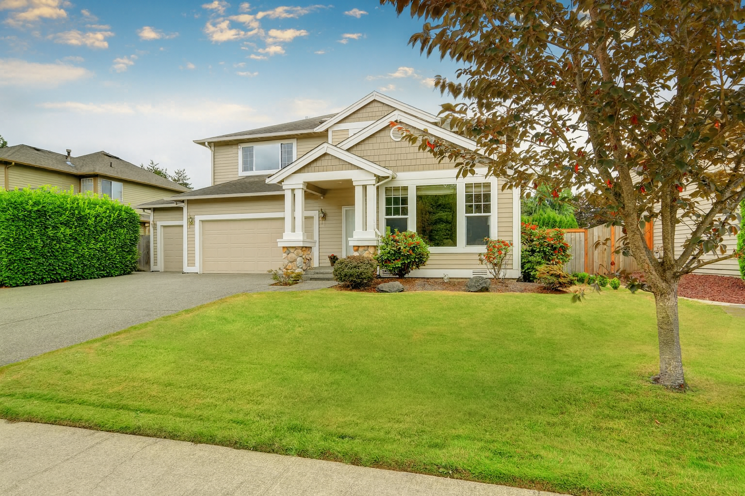 Two-story beige suburban home with white trim, stone accents, and a double garage. A neatly landscaped front yard with trees and shrubs complements the wide driveway and welcoming entryway.