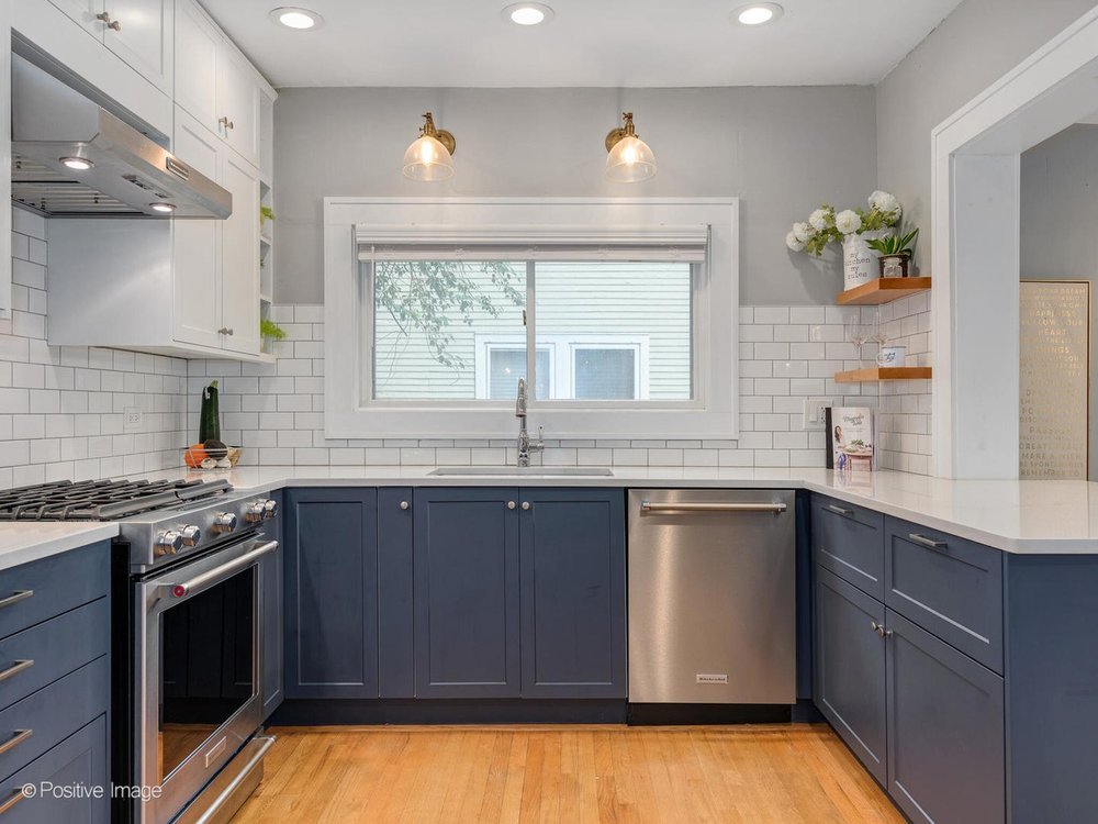 A bright, U-shaped kitchen featuring navy blue lower cabinets and white upper cabinets with shaker fronts. The design includes a subway tile backsplash, stainless steel appliances, and quartz countertops. Warm wood flooring adds contrast to the clean, modern palette. Two brass wall sconces provide accent lighting above a large window centered over the sink.