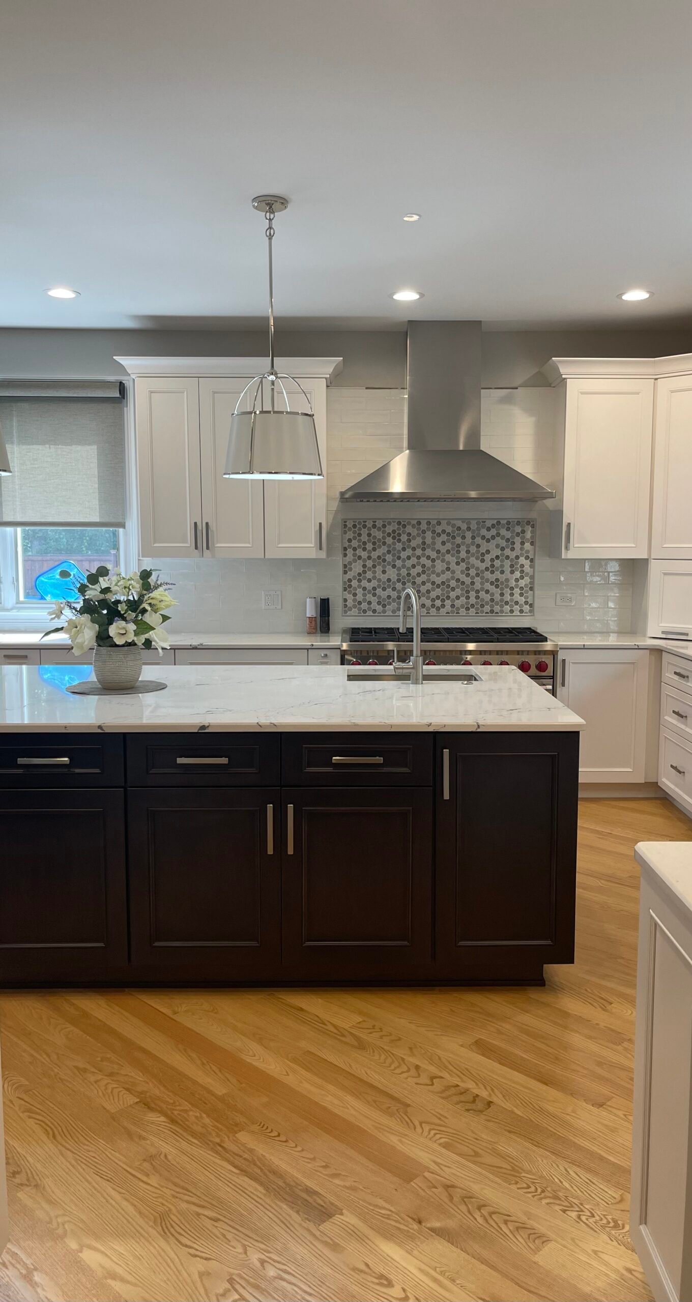 This Skokie kitchen remodel highlights a transitional design with a bold center island in rich espresso tones, contrasted against crisp white cabinetry. A stainless steel vent hood, geometric mosaic backsplash, and polished nickel hardware add sophistication, while the floral centerpiece brings softness to the clean, bright look.