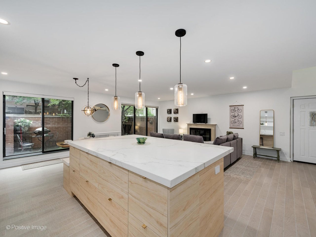 Wide view of a South Loop kitchen remodel with an open-concept layout flowing into the living room. Features include a large center island, pendant lighting, light wood cabinetry, and seamless connection to the family room and outdoor patio.